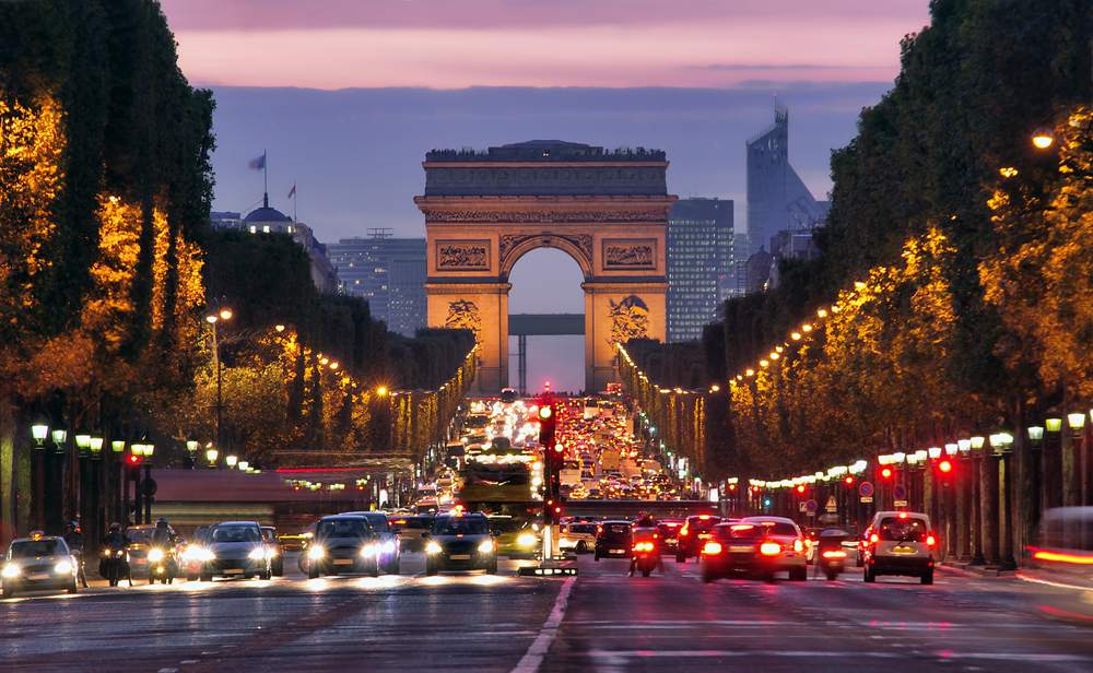 Dusk over the Champs-Elysees street with cars and lined with trees and the Arc de Triomphe at the end.