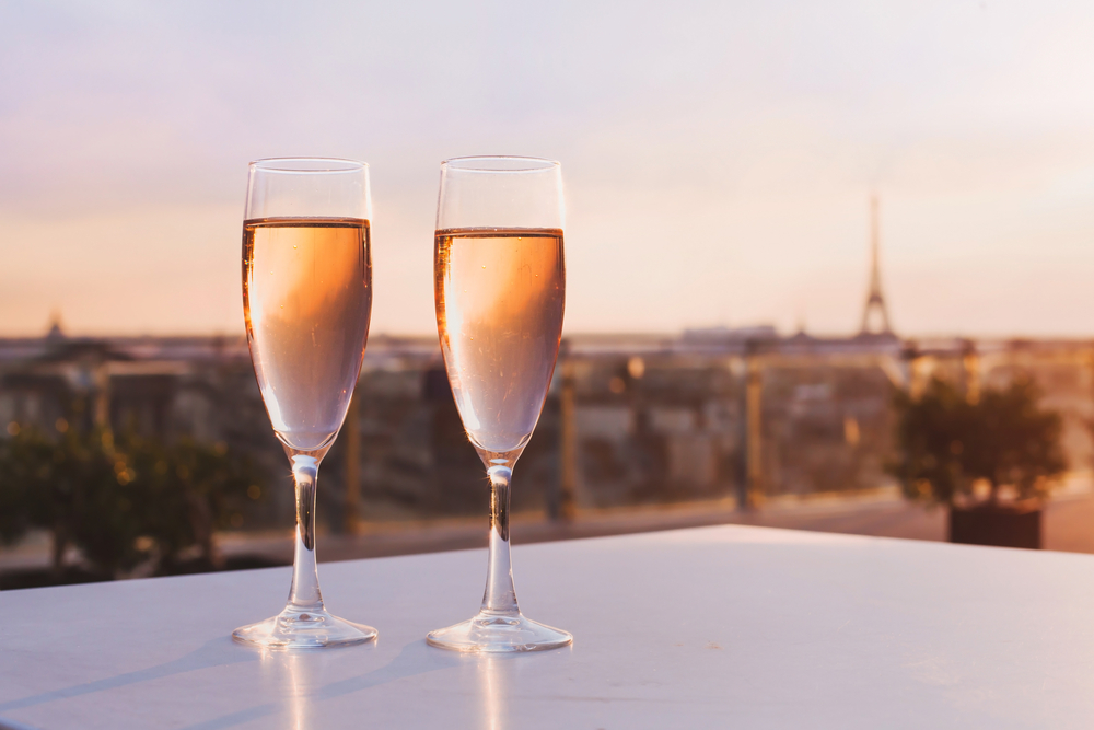 Pink sunset over two champagne flutes on a white table at a rooftop bar with the Eiffel Tower in the background.