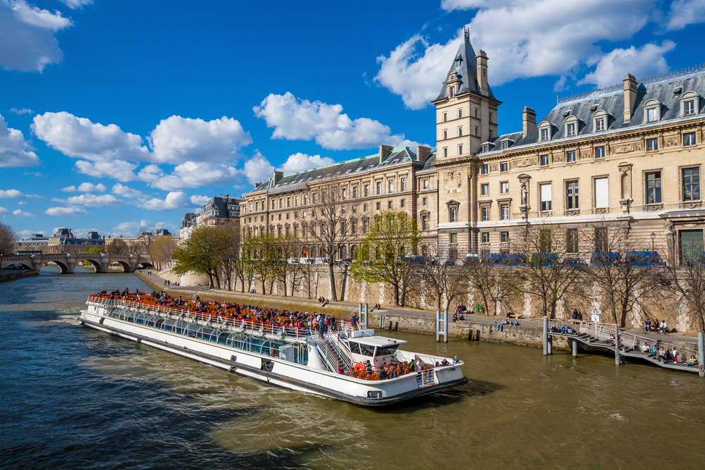 Tourists on an open deck of a cruise boat on the Seine River going by historic buildings.