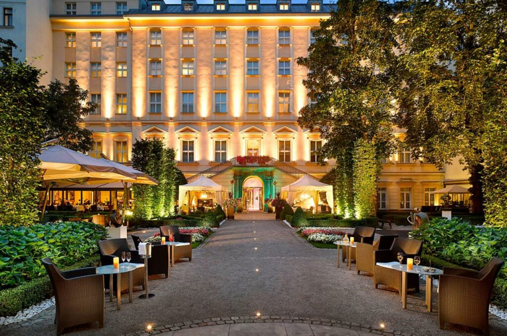 Garden with seating in front of the lit up Grand Mark Prague hotel at dusk.