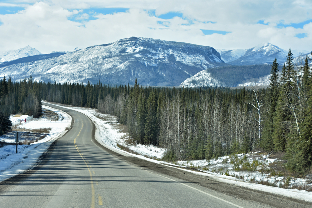 Alaska Highway in April. The road is clear but you can see trees either side and snowy mountains in the background. 