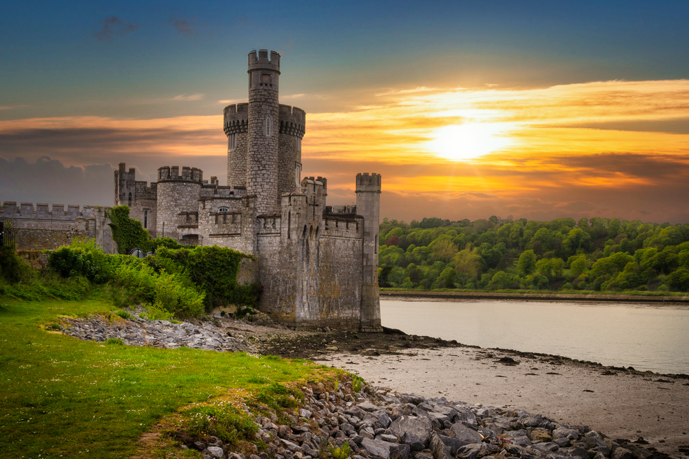 castle beside a river surrounded by greenery while the sun sets traveling to ireland