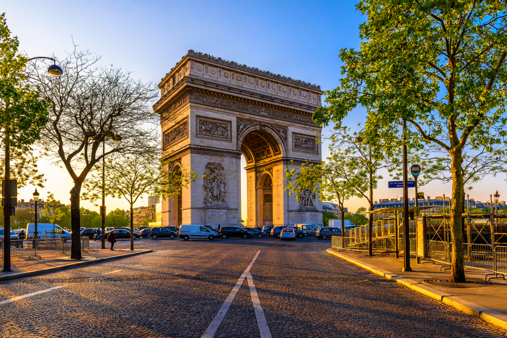 Late golden hour at the Arc de Triomphe with cars driving around it and trees in the foreground during 2 days in Paris.