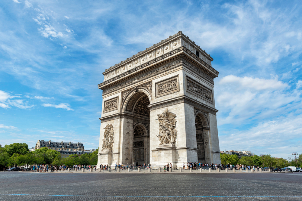 The towering, white Arc de Triomphe on a roundabout with many people walking below it during one day in Paris.