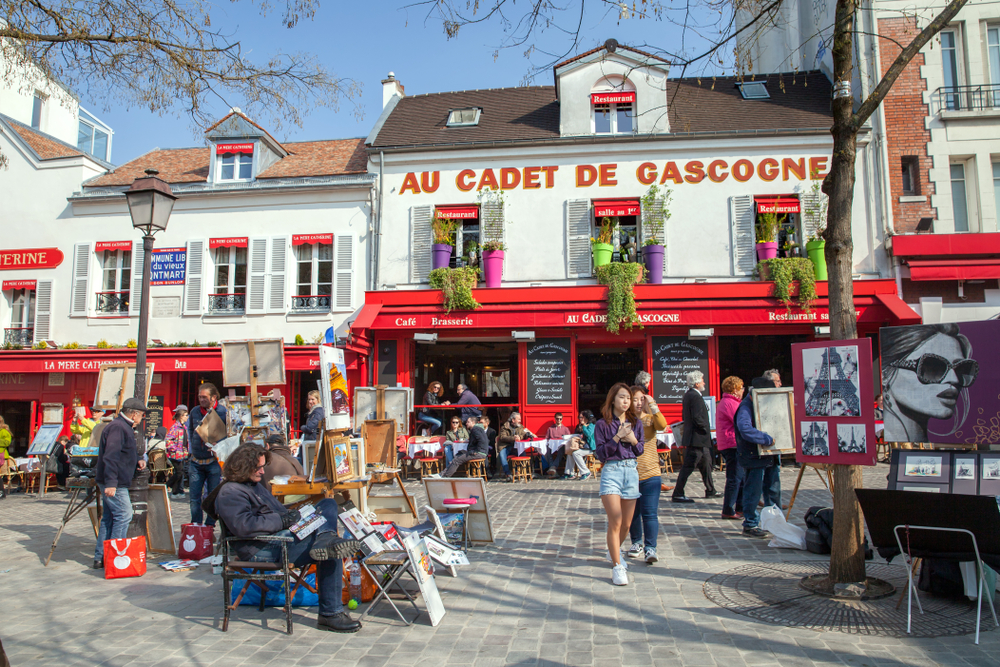Sunny day at Place du Tertre with many paintings and artworks on display and people walking around and a red and white cafe in the background.