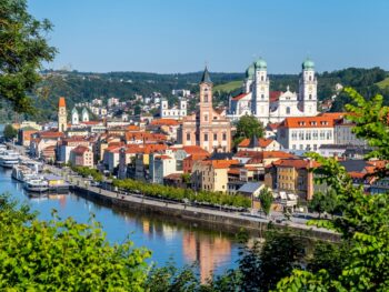 Sunny day over the river and colorful buildings of Passau, Germany, with many churches and tress in the foreground.