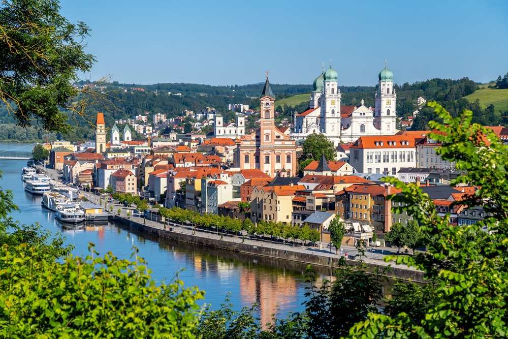 Sunny day over the river and colorful buildings of Passau, Germany, with many churches and tress in the foreground.