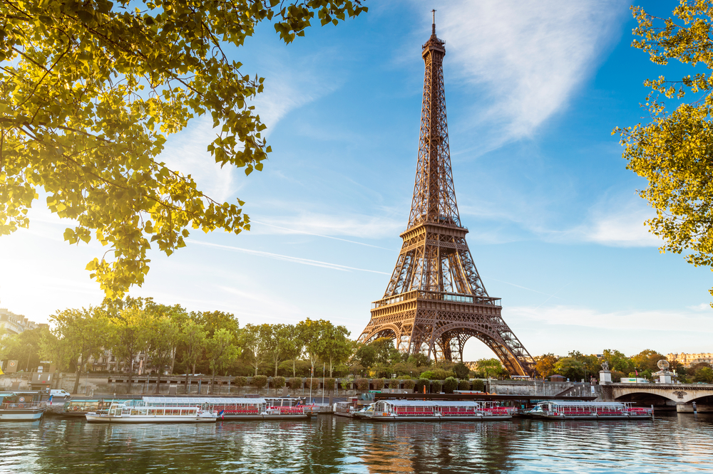 Golden hour view of the Eiffel Tower across the river with boats and trees in the foreground on a London-Paris itinerary.
