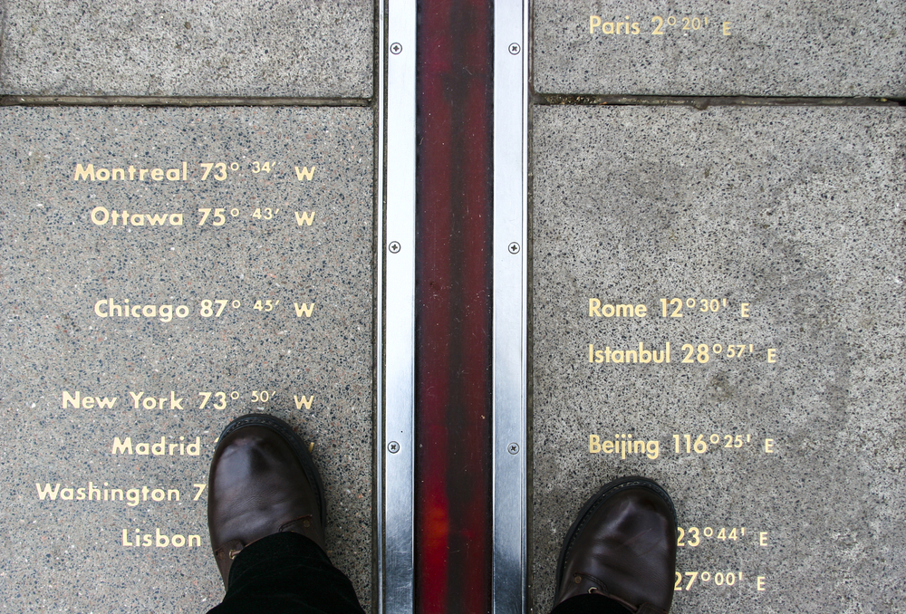 Looking straight down at shoes standing on the Greenwich Meridian with a list of different cities on each side.