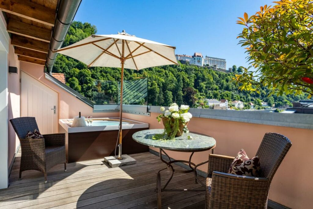 Terrace at Hotel Residenz Passau with wicker seats, table, umbrella, and a hot tub overlooking the city.