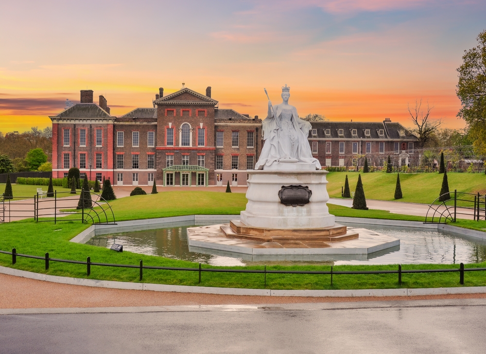 Vivid sunset over Kensington Palace with a white statue of Queen Victoria in front surrounded by a pool.