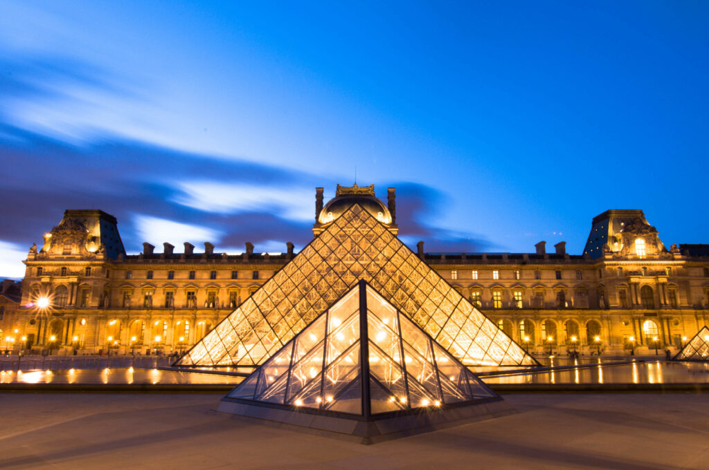 Dusk over the lit-up glass pyramids with the Louvre Palace behind them.