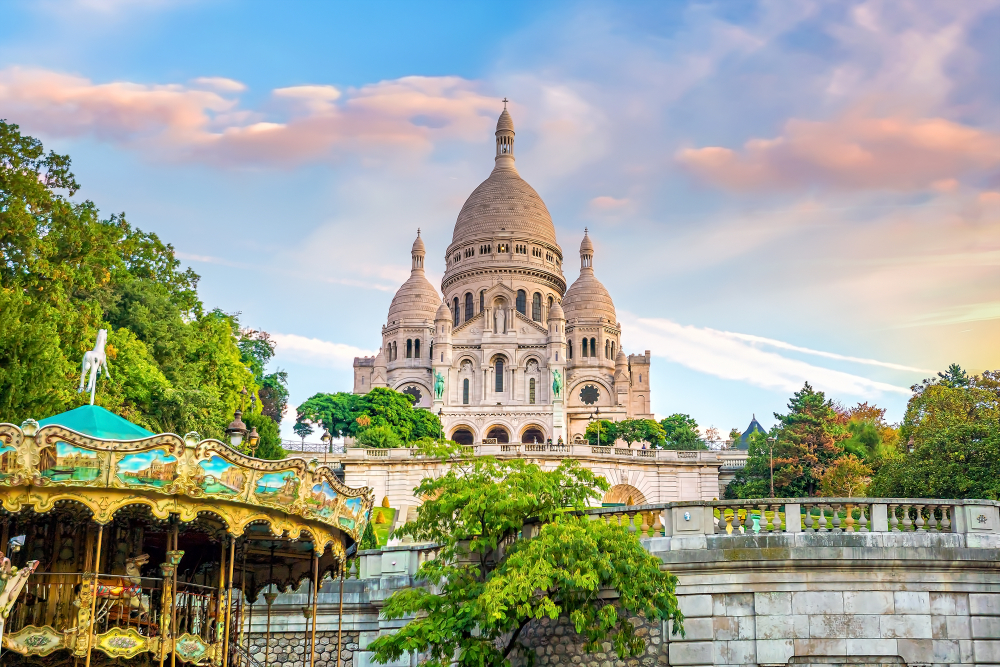 Looking up at the white, domed Sacre-Coeur Basilica with a merry-go-round in the foreground under a pastel sunset.