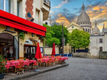 Red chairs and tables outside of a cafe in Montmartre with the white Sacre-Couer Basilica in the background at sunset.