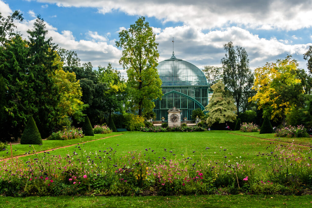 Glass conservatory in center of picture surrounded by lush green trees and green grass. Colorful flowers in foreground.