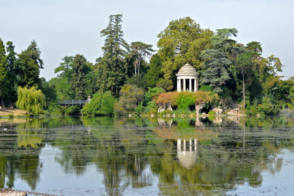 Fancy round building surrounded by trees and greenery with water in foreground. Gardens in Paris