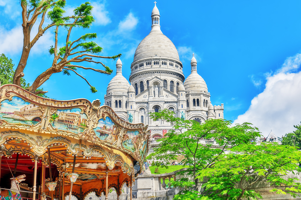 Looking up at the beautiful, white Sacre-Coeur Basilica with three domes and a merry-go-round and trees in the foreground.
