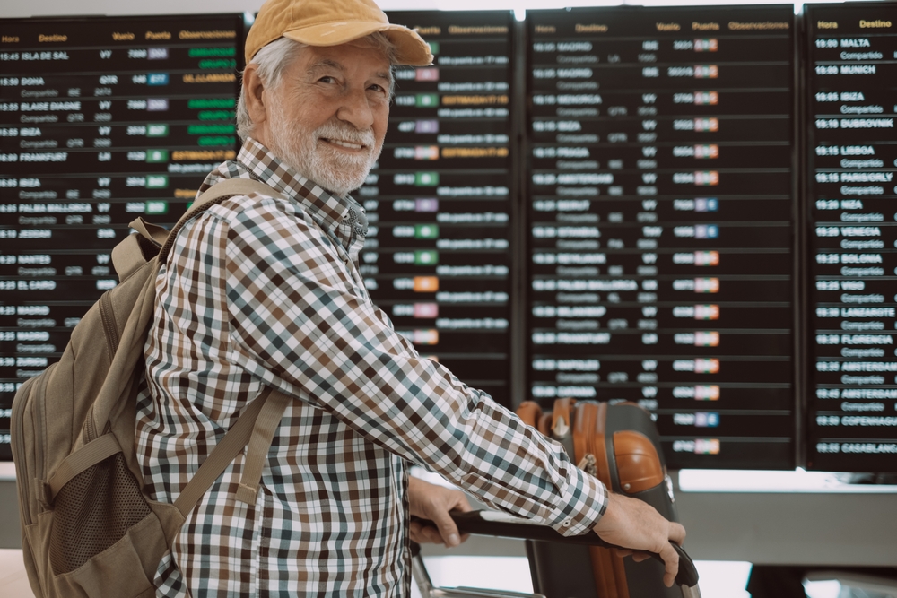 elderly man in front of airline departures board with grey beard and brown checked shirt