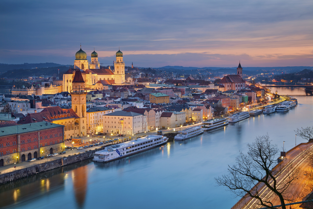 Purple dusk over the town of Passau, Germany, with cruise ships docked on the river, street lights glowing, and churches lit up.