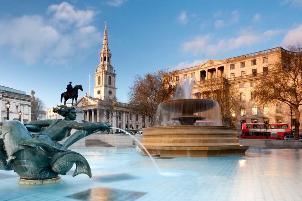 A fountain with a statue in Trafalgar Square with a church in the background.