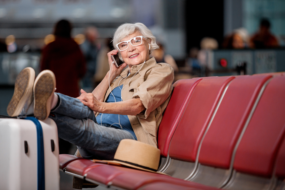 Solo senior travel tips show grey haired woman sitting on red couch talking on phone
