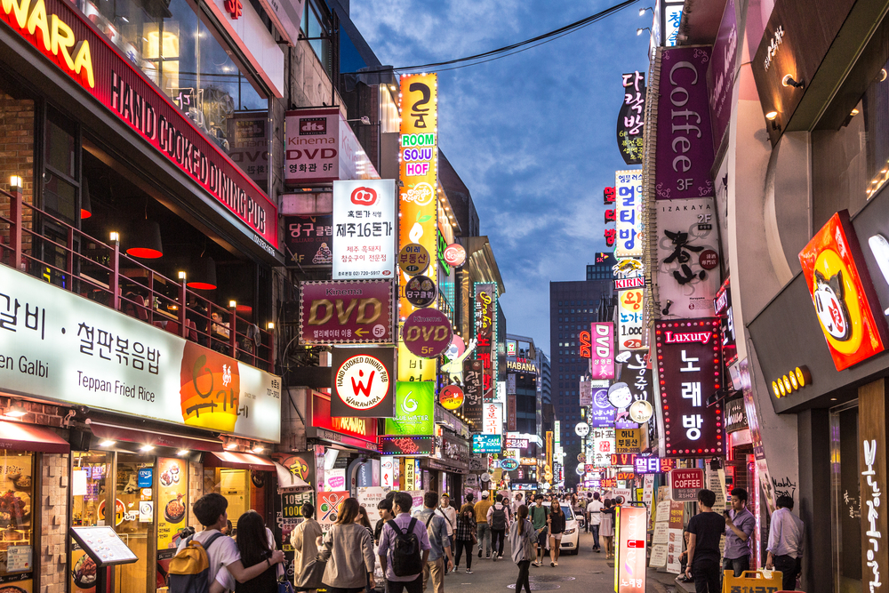 Nighttime on a busy street with people walking around and many bright colorful signs in Korean and some English.