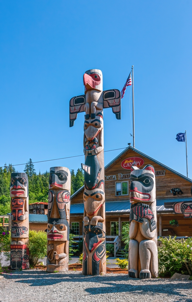  4 colorful totem poles in front of brown building under blue sky. Green foliage around.