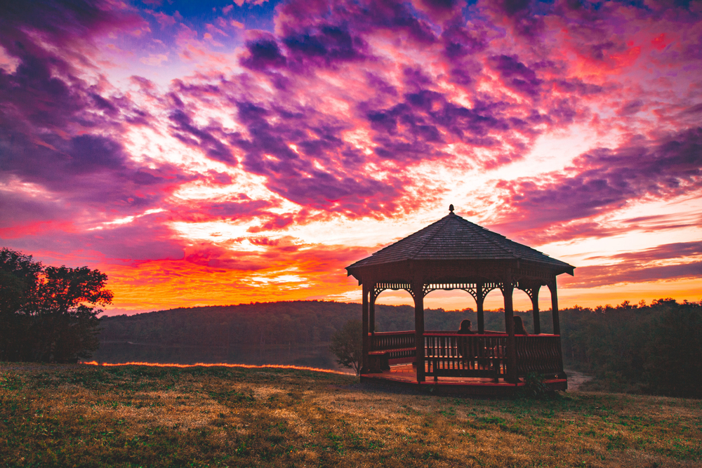 Sunrise over gazebo in front of lake in Pocono mountain. Couple are sat together. Article is about romantic getaways in the Poconos.