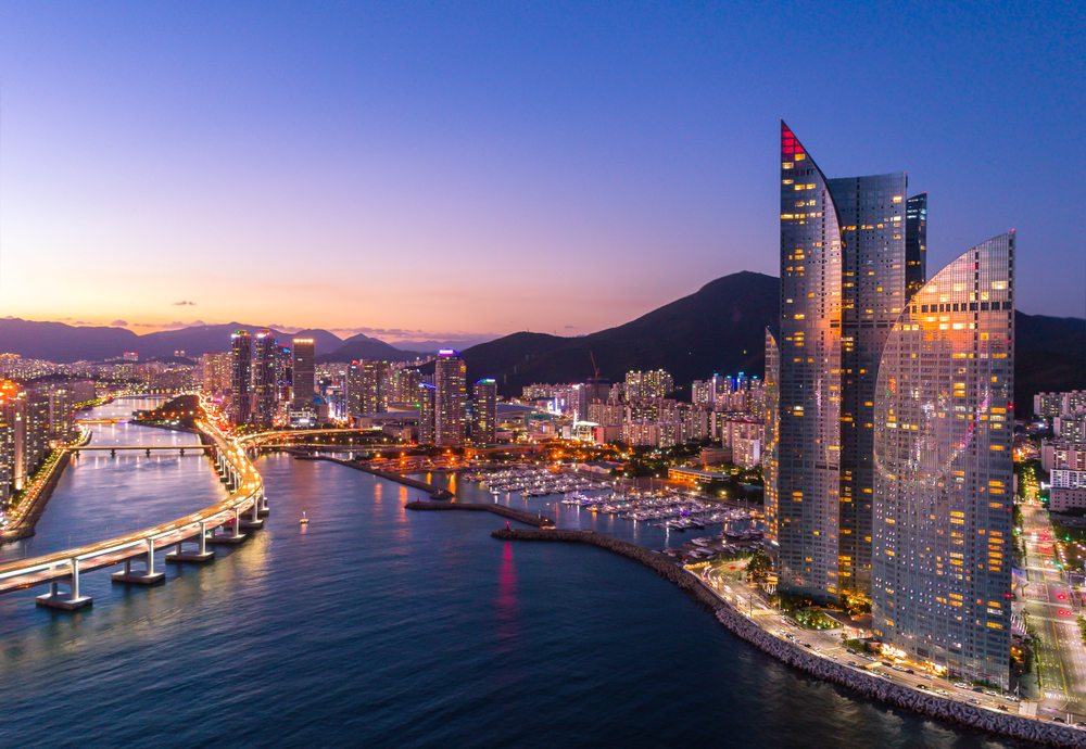 Purple twilight over the harbor and skyscrapers of Busan, South Korea, with mountains in the distance.