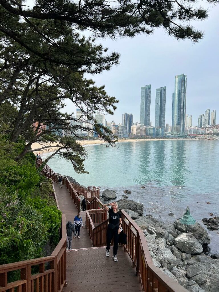 Older, blonde woman posing on the Dongbaek Island Coastal Promenade with the beach and skyline in the background on a cloudy day.