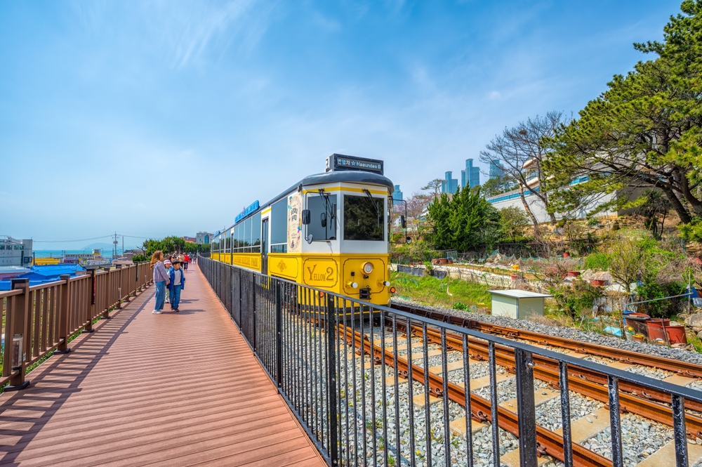 People walking along the Busan Green Railway Coastal Walking Trail next to a yellow train.