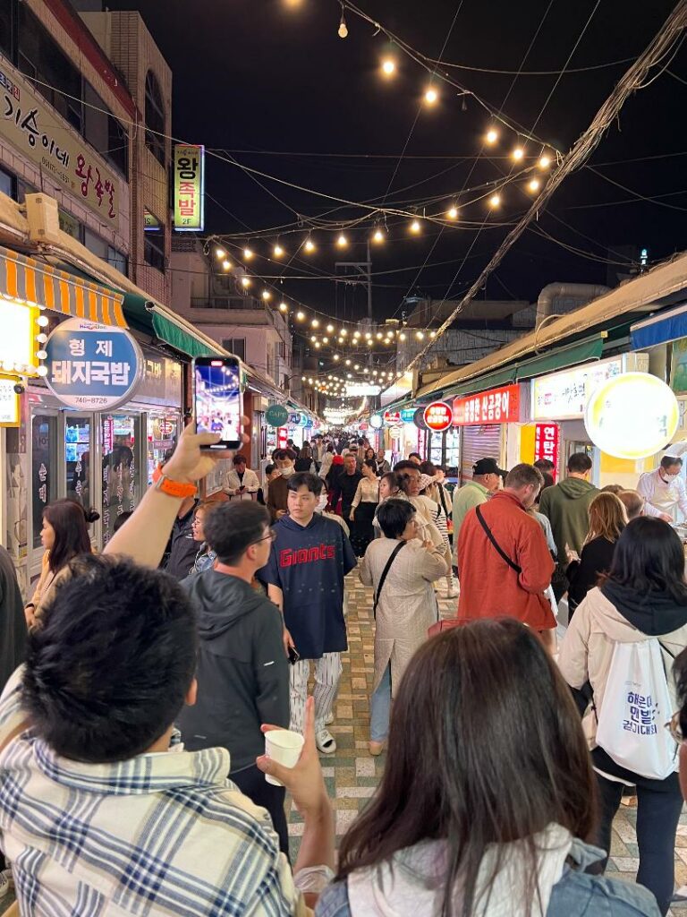 Nighttime at the Haeundae Traditional Market with strung lights overhead and many people walking among lit up food stalls.