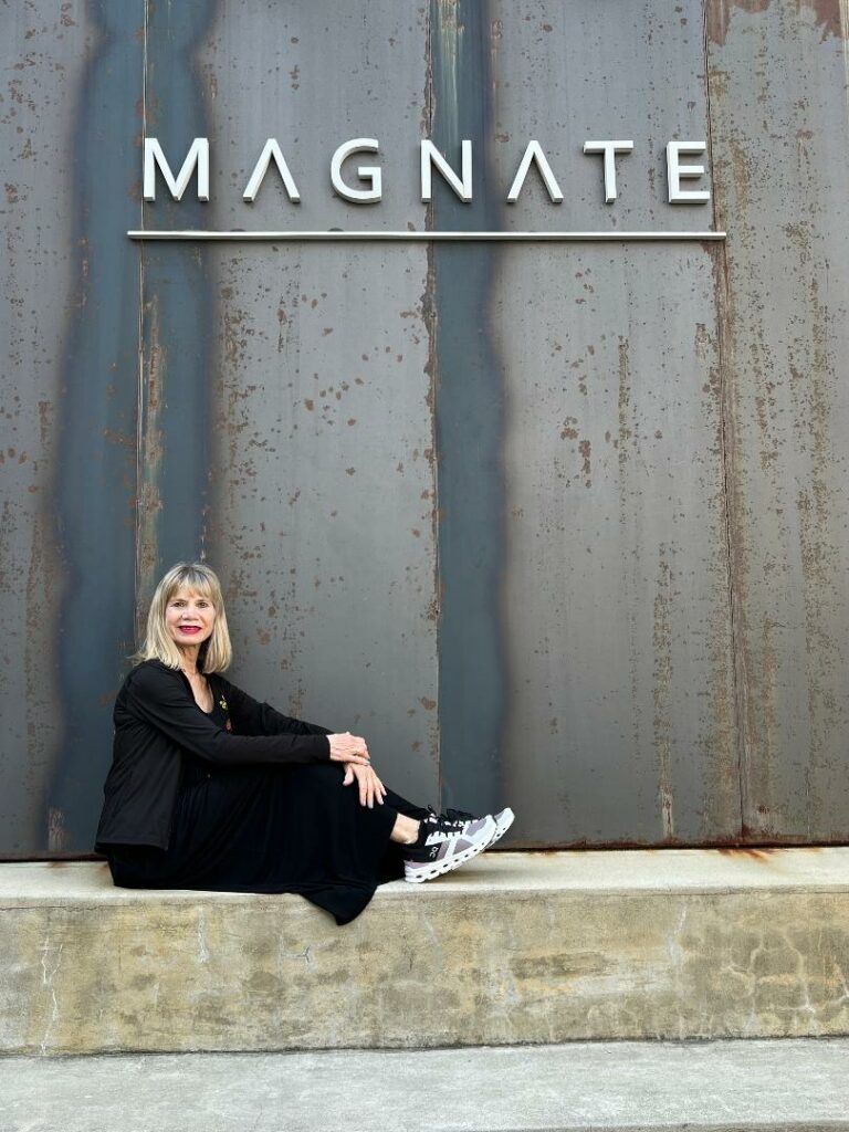 Older, blonde woman wearing all black sitting in front of the Magnate Cafe sign.