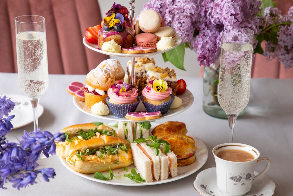 Traditional English afternoon tea with selection of cakes and sandwiches. There are champagne glasses on the table and purple flowers in the background. 