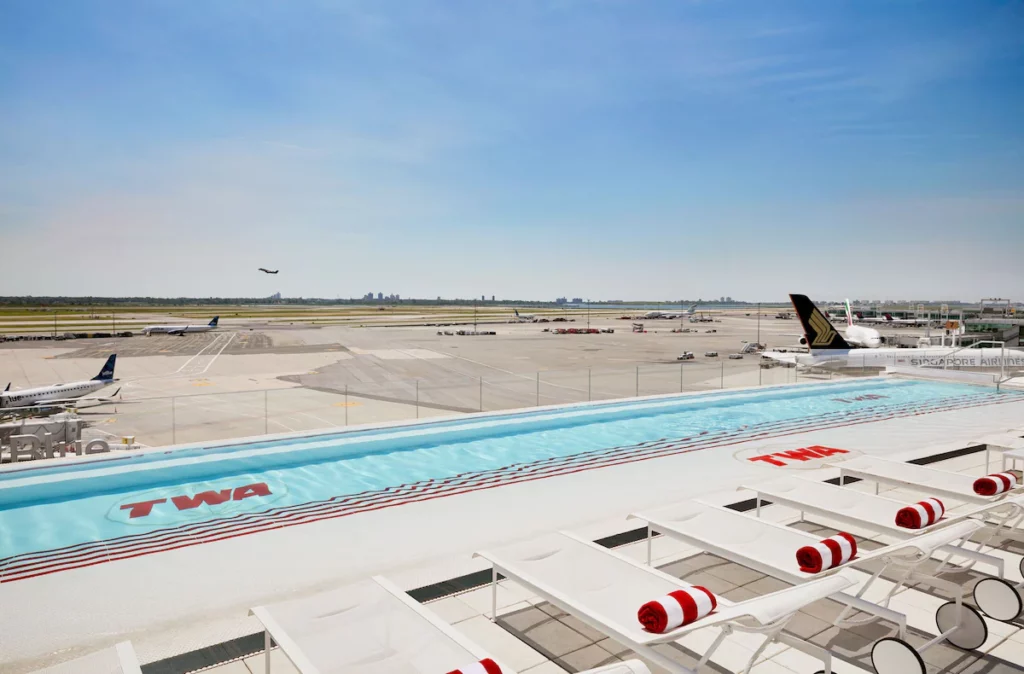 The swimming pool at the TWA Hotel  looks over the runway. It has white loungers and red and white towels. 