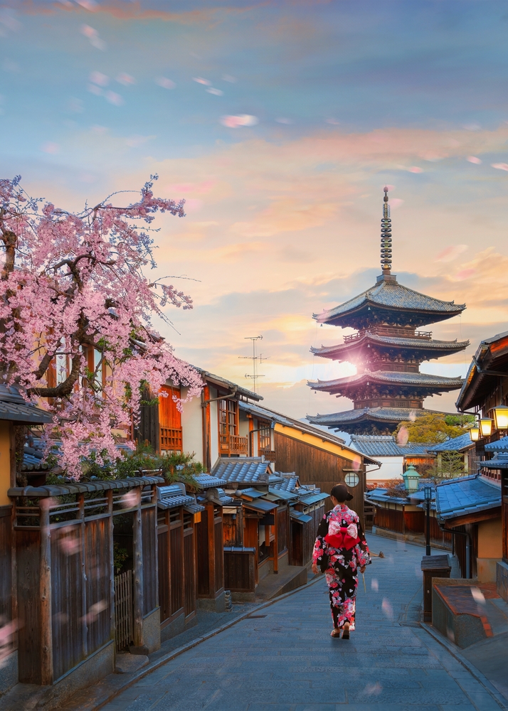 Scenic cityscape of Yasaka pagoda sunset in Kyoto with a young Japanese woman in a traditional Kimono dress during full bloom cherry blossom and scatter sakura petals.