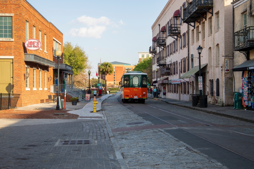 An orange and green trolley bus from Old Town Trolley driving on River Street with shops and restaurants and people walking along the cobblestone sidewalk in Savannah