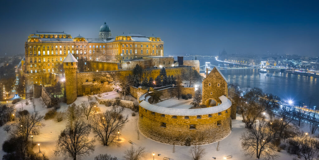 Aerial view of illuminated Buda Castle Royal Palace on a winter night with snowy park and Szechenyi Chain Bridge in Budapest Hungry one of the places to visit in Europe in winter. 