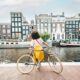 girl with cycle in front of canal with buildings and boats in the backdrop