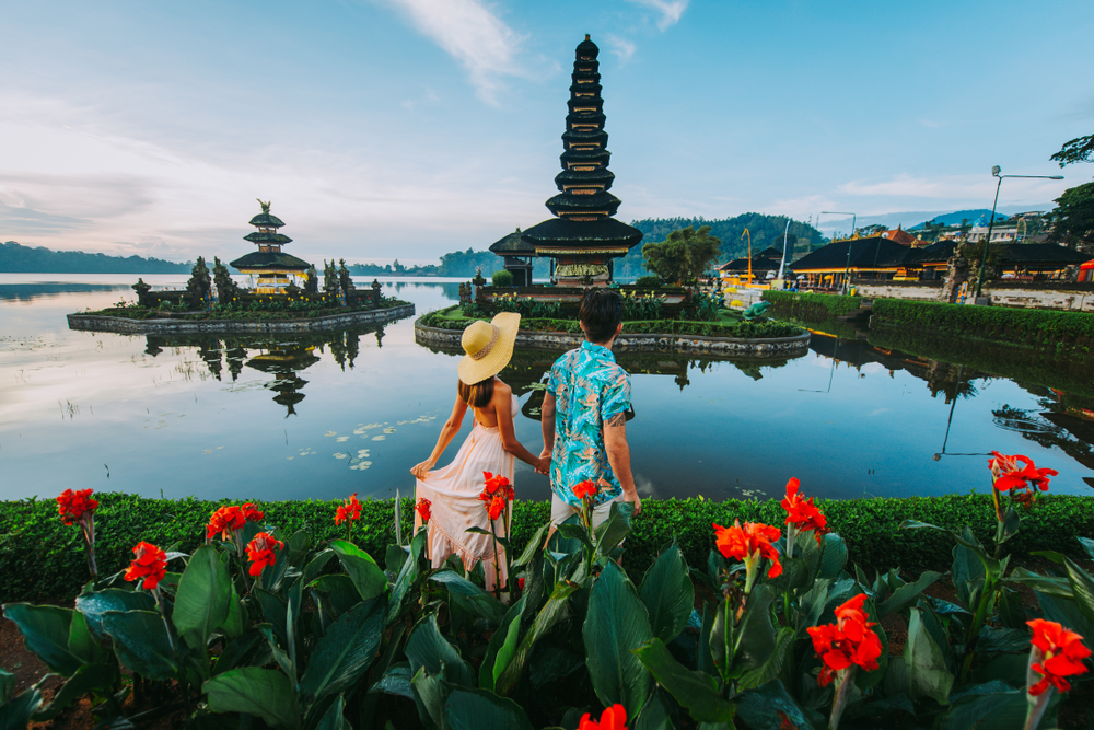 A couple holding hands and walking along the water near red flowers at the Ulun Danu Beratan Temple in Bali.