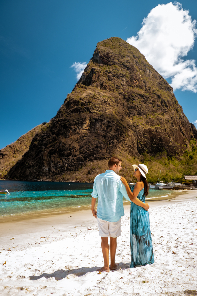 A couple standing on a white sand beach with a tall hill in the background in St. Lucia, one of the best honeymoon destinations.