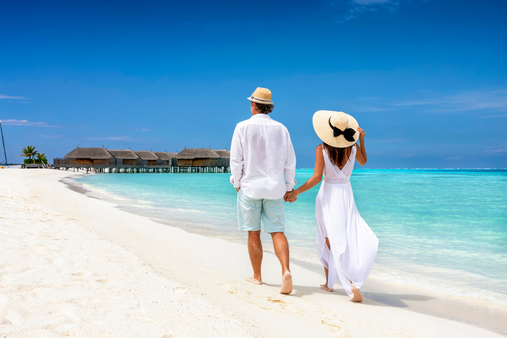 A couple in white walking along the white sand beach in the Maldives heading towards overwater bungalows.