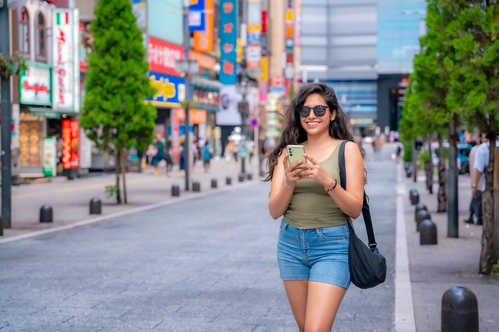 Woman in a tank top, shorts, and sunglasses walks down a street while texting on a phone.