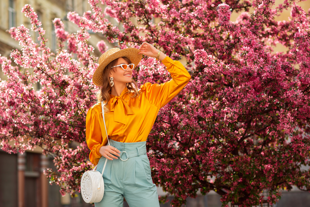 Happy smiling woman wearing trendy straw hat, yellow satin blouse, blue trousers, cat eye sunglasses, with wicker bag, posing in street near pink spring blossom trees