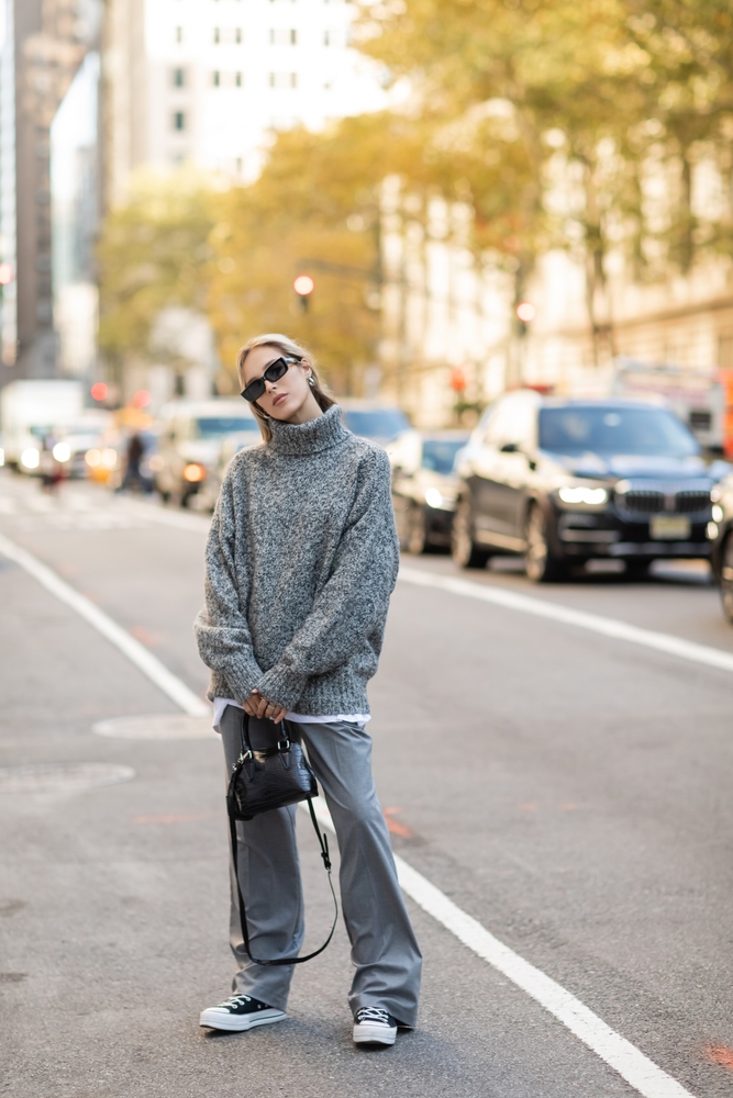 full length of stylish woman in sunglasses and grey outfit holding black handbag on street of New York city