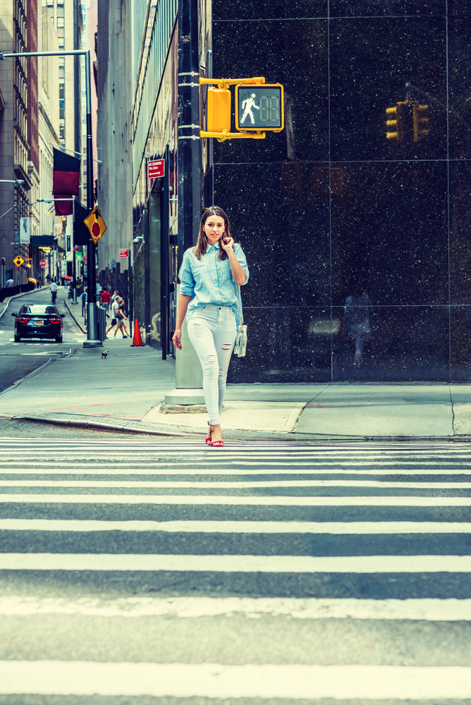 Young Woman wearing light blue striped shirt, white destroyed jeans, red sandal heels, shoulder carrying small bag, walking across street in New York. 