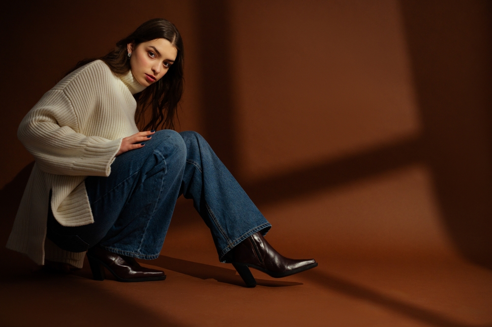 woman wearing oversized white sweater, wide leg jeans, leather cowboy boots, posing on brown background. Article is about what to wear in NYC 