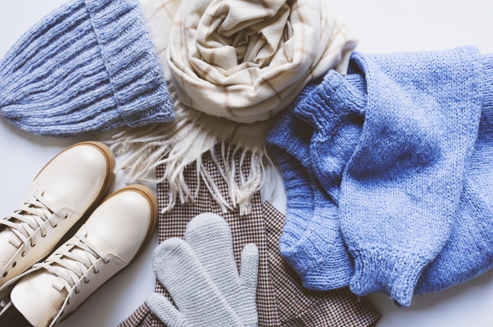 Flay lay image of winter clothes, including a blue sweater, beanie, scarf, gloves, and shoes.