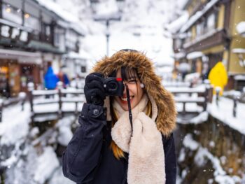 Woman holding a camera to her face wearing a hooded coat and fluffy scarf, one of the best outfits in Japan for Winter. In the background is a snowy, small town in Japan.