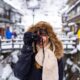 Woman holding a camera to her face wearing a hooded coat and fluffy scarf, one of the best outfits in Japan for Winter. In the background is a snowy, small town in Japan.